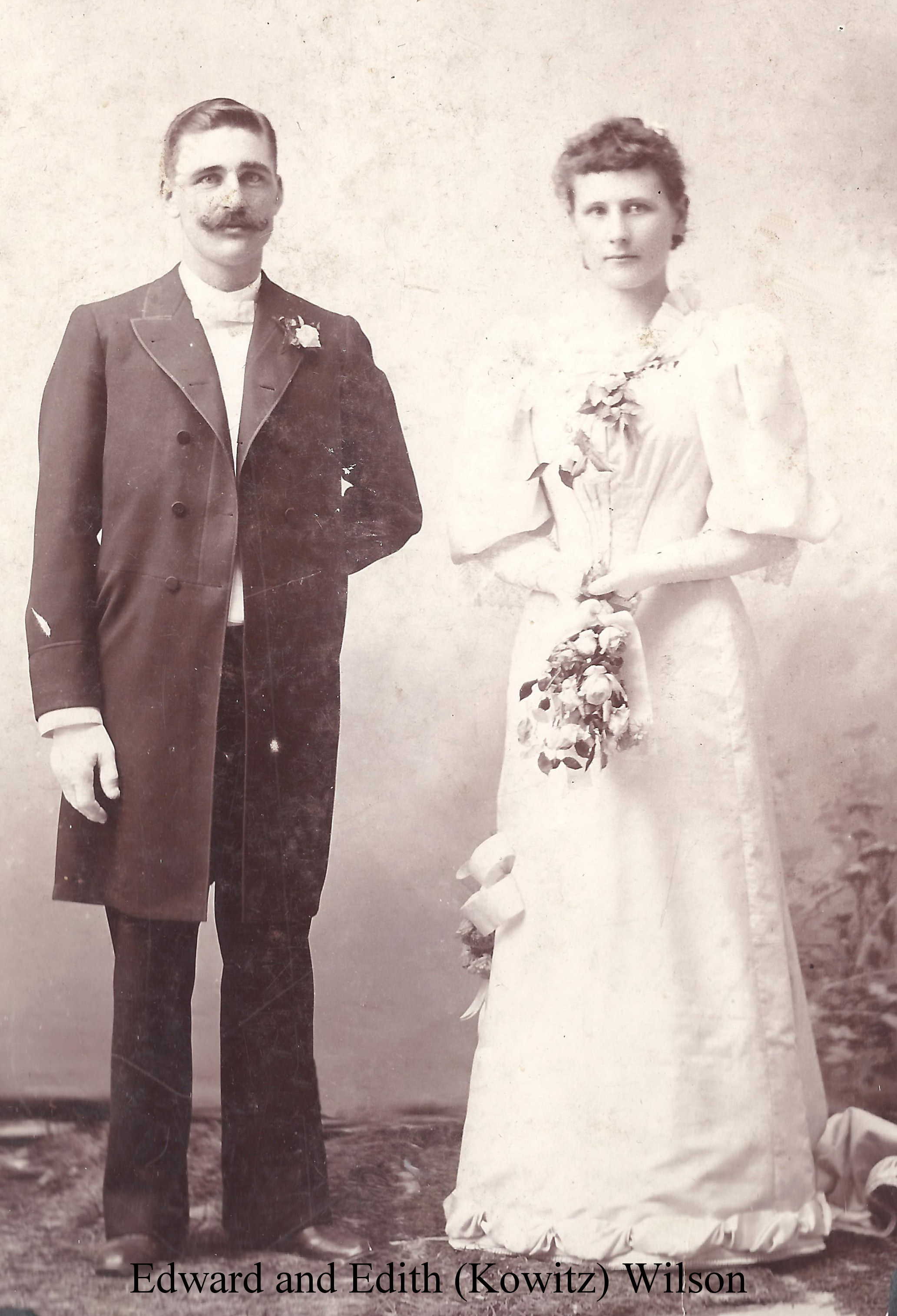 IMAGE/PHOTO: Edward and Edith (Kowitz) Wilson: Black and white wedding photograph of Ed Wilson at left, wearing a knee-length coat with a boutionniere,with white tie and gloves, his left hand inserted into the coat at his waist, and Edith Kowitz, in a white floor-length dress with elbow-length flounce sleeves and flowers at the neck and at her right knee, holding a bouquet low; they both stare unsmiling at the camera.